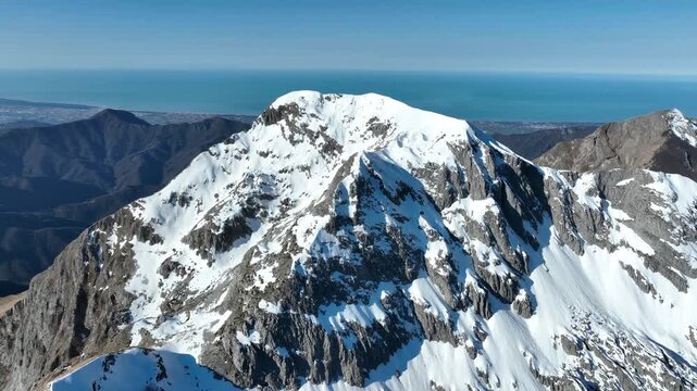 Aerial view of the snow-covered Pania della Croce mountain peak with rocky slopes and a distant view of the coastline in Stazzema, Tuscany, Italy.
