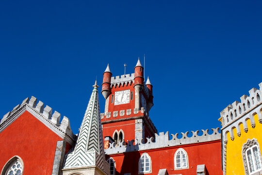 Sintra, Portugal - 18 August 2017: View of the colorful Pena Palace with its distinctive red and yellow walls, ornate clock tower, and intricate architectural details against blue sky.