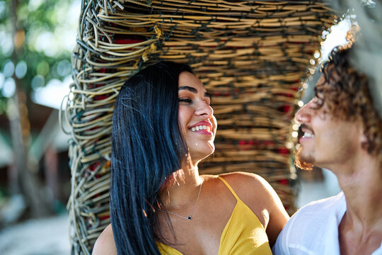 A smiling couple shares fresh coconuts while relaxing inside a woven beach nest on a sunlit tropical shore. The woman in a yellow dress and her partner laugh together, capturing a carefree summer vaca
