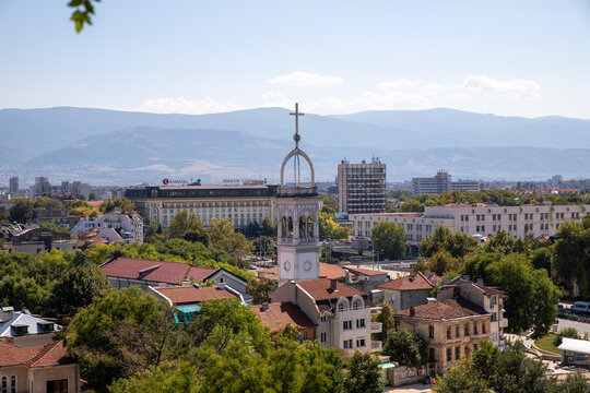 Plovdiv, Bulgaria - 24 August 2018: View of the historic church tower rising above the rooftops and lush trees in the city center with mountains in the background.