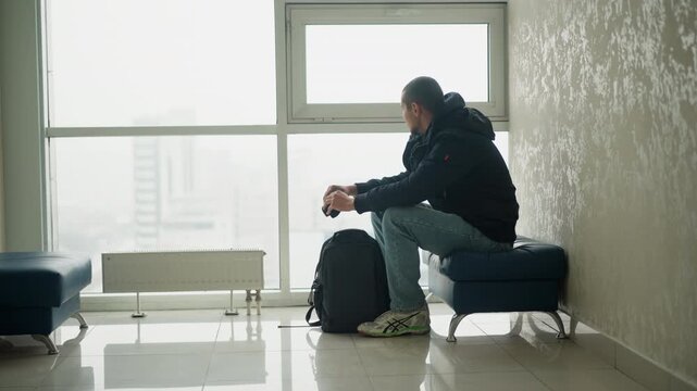 Man sits on blue bench in indoor waiting area holding small objects with minimal movement. Man wears casual clothing and has luggage beside him as man looks out large windows showing city
