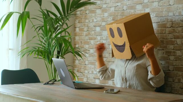 Happy businesswoman wearing a cardboard box with a smiling emoticon dancing and celebrating success in her modern office after finishing work on the laptop, symbolizing happiness at work
