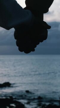 Close-up silhouette of a young couple holding hands with a calm sea and cloudy sky in the background, a beautiful and poignant moment of love, support, and togetherness