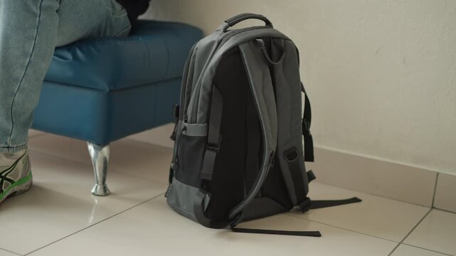 Close up of gray backpack on floor next to blue furniture with part of person wearing jeans and sneakers visible. scene is indoors featuring white tiled flooring and plain wall background