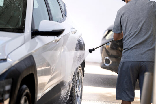 Man cleaning white automobile with water spray at a public car wash facility