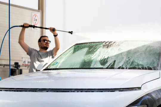 Man washing vehicle windshield with high-pressure hose, soap suds covering glass