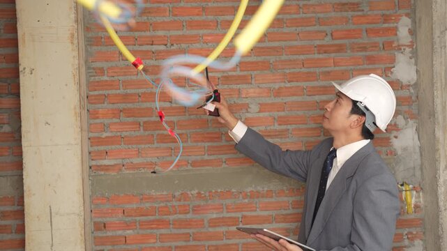Construction engineer inspecting electrical wiring on unfinished brick wall, electrical installation, building inspection, site safety, quality control, construction engineering.