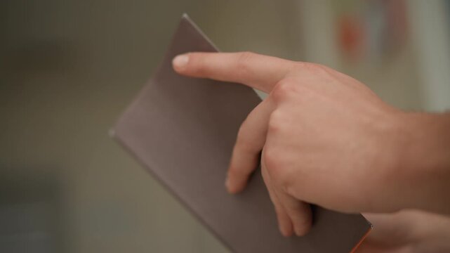 Close up of hand holding and pointing at corner of closed notebook with soft lighting and shallow depth of field. scene focuses on human interaction with stationery in indoor environment