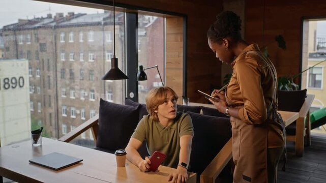 African American waitress taking order with digital tablet from seated Caucasian young adult customer in cafe. Ideal for hospitality service promotions, POS software marketing, or staff training