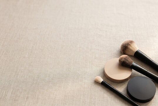 Various makeup tools are arranged neatly on a light fabric surface. Three brushes and two compacts are visible, showing their colors and styles