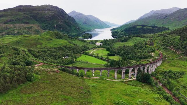 Aerial view of Glenfinnan Viaduct on the West Highland Line, with Loch Shiel and a green Scottish Highlands valley under cloudy skies in Scotland, United Kingdom. 4K UHD video.