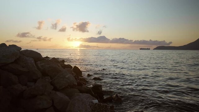 A vibrant summer sunset over the calm sea, with an orange-red sky glowing above large coastal stones and its warm reflection shimmering across the water.