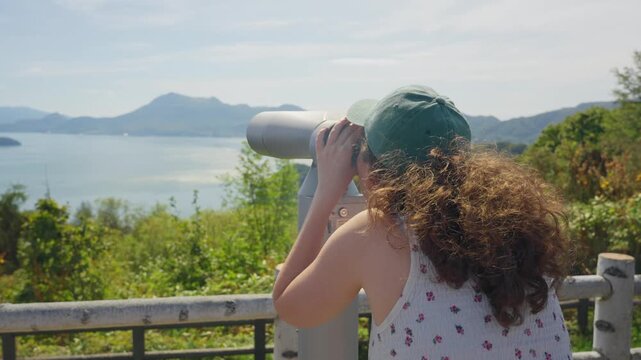 Young tourist with long curly hair and a green hat looking through coin operated binoculars at the beautiful scenic landscape of Lake Toya on the island of Hokkaido in Japan during autumn season