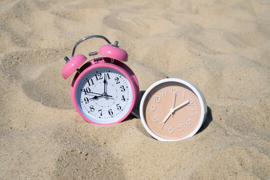 Pink retro alarm clock and modern beige clock on beach sand for time zones travel countdown and vacation concept	
