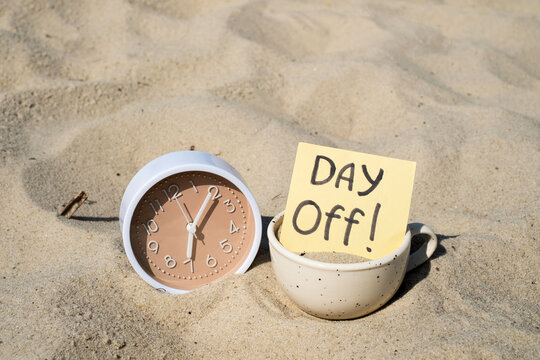 Beige clock and coffee cup with DAY Off note sticked in beach sand for vacation time and work life balance concept
