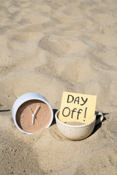 Beige clock and coffee cup with DAY Off note sticked in beach sand for vacation time and work life balance concept	