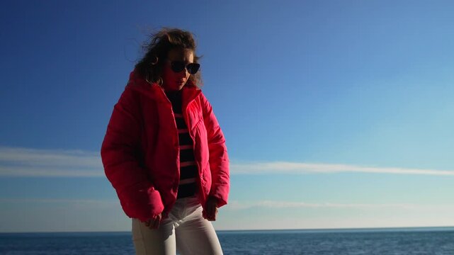 Woman beach ocean stands on the shore during a bright sunny day wearing a vibrant pink puffer jacket and sunglasses