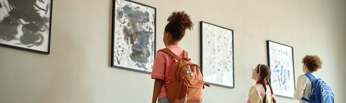 Diverse children with backpacks looking at paintings on wall in art gallery, rear view. Image suitable for school field trip promotions and cultural education campaigns
