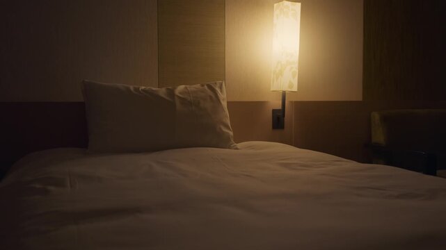 Dimly lit and empty hotel bedroom with a bedside lamp casting a warm glow on the neatly made bed, pillow, and headboard. A cozy and tranquil ambiance for a relaxing stay. Mysterious dark motel room