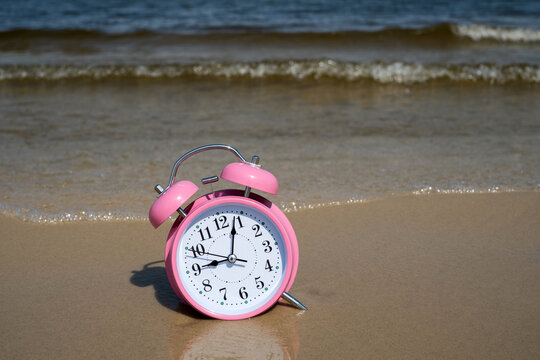 Pink alarm clock standing on wet sand beach near ocean waves for summer vacation time concept	