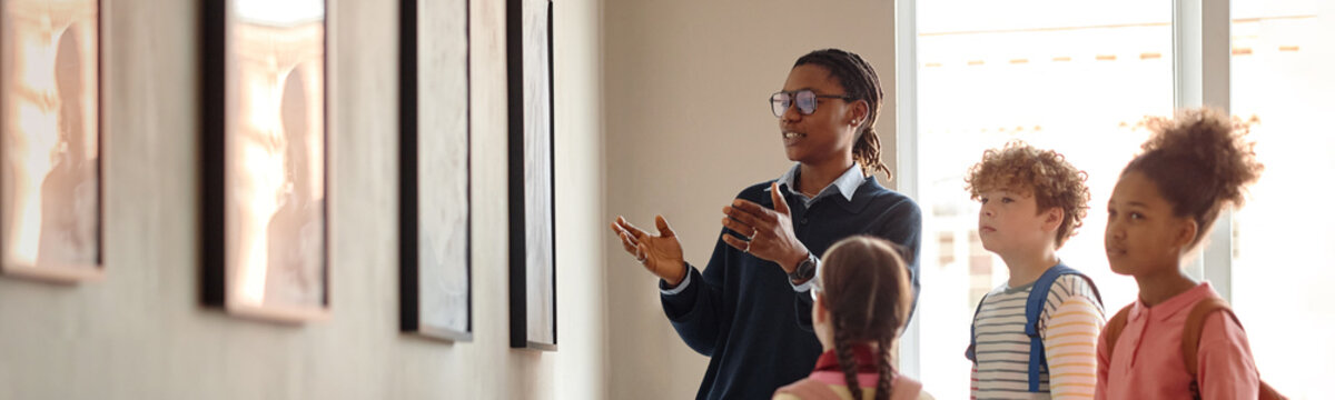 African American male guide explaining paintings to diverse group of children in art gallery, promoting cultural education and school field trips, medium shot