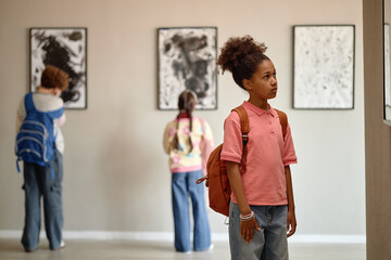Fototapeta premium African American schoolgirl looking at paintings in art gallery during field trip with classmates. Image suitable for promoting museum education programs and youth cultural activities