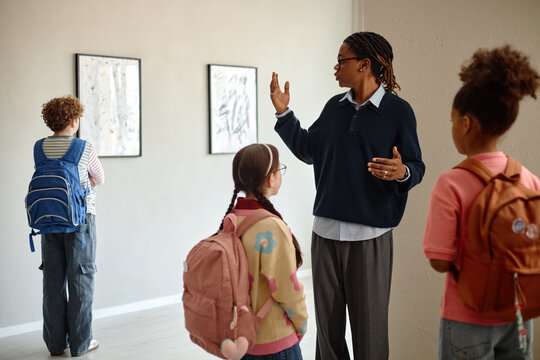 African American male teacher guiding diverse group of school children in art gallery, explaining paintings. Image suitable for educational programs and museum field trip promotions