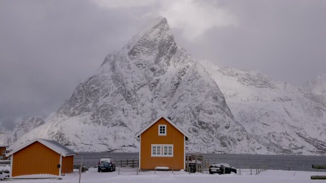 View of yellow cabins set against a dramatic snow-covered mountain in the winter landscape of Hamnoy, Nordland, Norway.