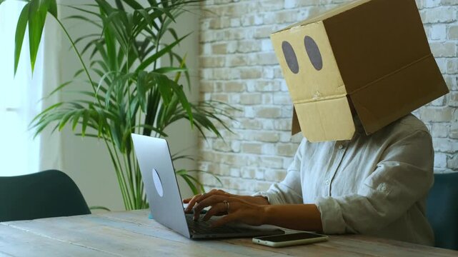 Unrecognizable person with a box on her head working remotely on a laptop computer, typing on the keyboard. Concept of employee alienation, modern work life, and online identity