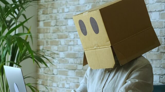 Funny character with a cardboard box on their head working on a laptop at a modern office desk, symbolizing anonymity in the digital age, introversion, or hiding one's identity