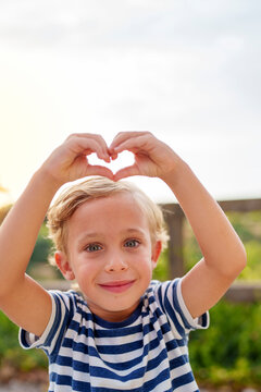 Little boy forming heart shape with hands, expressing love, care, and happiness outdoors looking at camera