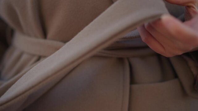Close up of hands adjusting brown coat belt and fabric showing texture detail as subject secures their outerwear. focus remains on hand movement over soft lighting with no background