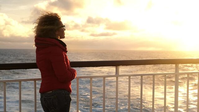 Pensive woman with curly hair wearing a red jacket, standing on the deck of a moving ferry and enjoying the beautiful sunset over the sea while her hair blows in the wind