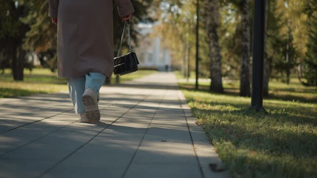 Close up of woman walking down sunlit park pathway holding small black handbag. Woman wears light blue jeans and beige shoes under long brown coat with trees lining path in soft daylight
