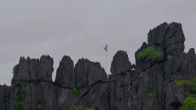 Slow right-to-left aerial drift past jagged tropical rock formations in the rain, with the New Caledonia Kanaky flag waving along the ridge beneath a gray sky.