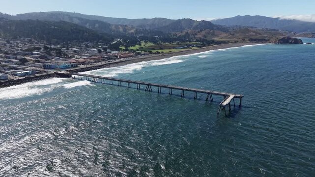 Scenic aerial view of the Pacifica Municipal Pier stretching into the Pacific Ocean, with gentle waves rolling onto the sandy shore of the California coast and a small town in the background