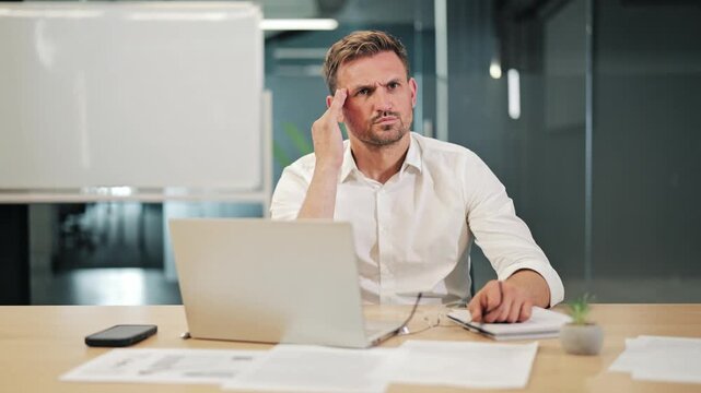 Businessman experiencing intense headache while working at laptop. Workplace pressure and overwork cause discomfort, seeking relief from pain and stress. Feeling unwell at office desk.