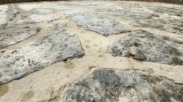 Large stone pavers stretch across a sunlit pathway. Fine sand fills the narrow gaps between the rough rocks. The rustic texture creates a sense of warmth, stability, and calm.