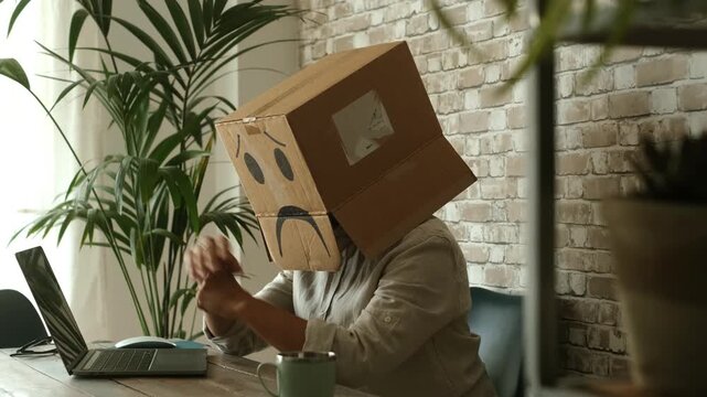 Employee with a sad face cardboard box on their head feeling desperate and stressed at work, showing professional burnout and fatigue from overworking while sitting at an office desk