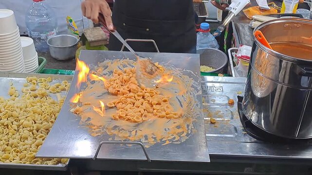Street food vendor torches creamy macaroni cheese on hot griddle with flames at night festival. Cheesy pasta, blowtorch cooking. PAMEKASAN, INDONESIA - JUNE 08, 2026