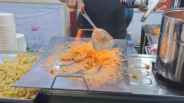Street food vendor torches creamy macaroni cheese on hot griddle with flames at night festival. Cheesy pasta, blowtorch cooking. PAMEKASAN, INDONESIA - JUNE 08, 2026