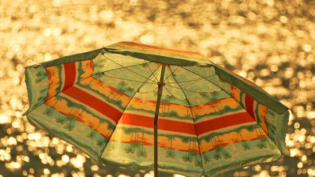 Colorful beach umbrella shields against golden sunset light reflecting off the sea. Fixed medium shot.