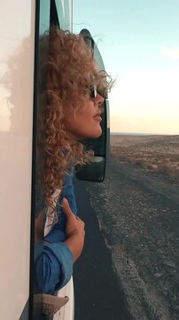 Carefree curly-haired woman with sunglasses feeling the wind in her hair while looking out of a camper van window, enjoying the freedom of a road trip through a desert landscape at sunset