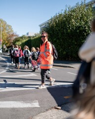 Fototapeta premium Hispanic crossing supervisor in a bright orange hi-vis coat guiding children across a sunny street, with copy space for road safety and back to school programs