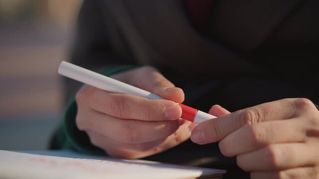 Close up of hands holding red and white marker over open notebook with faint text. person manipulates pen revealing its tip while interacting with paper in soft natural light. Background