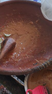 Close up of hands grinding Madura petis, peanuts, chili, tomato in clay mortar for authentic rujak cingur. Traditional Indonesian street food preparation, East Java culinary culture, spicy sauce.