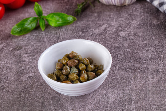 Pickled capers in white bowl on stone table with fresh basil and tomatoes