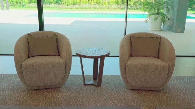 Elegant living room in a modern home featuring two designer armchairs and a small coffee table overlooking a beautiful backyard swimming pool and garden through a large panoramic window