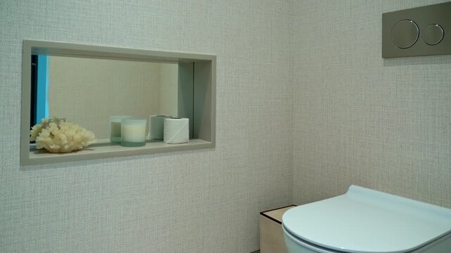 Modern minimalist bathroom interior with wall mounted toilet and recessed niche shelf displaying coral, candles and toilet paper against a textured white wall, bright and elegant