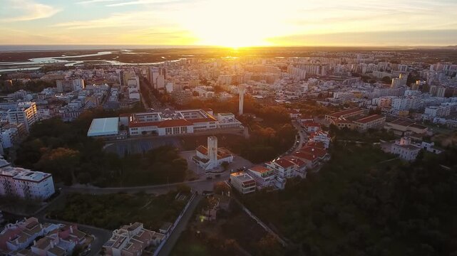 Golden sunset illuminating the city of faro, portugal, with an aerial perspective showcasing the church of santo antonio alto, the urban landscape, and the distant ria formosa natural park